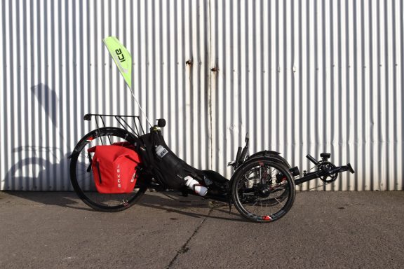 A recumbent tricycle with a red pannier and yellow flag against a corrugated backdrop.